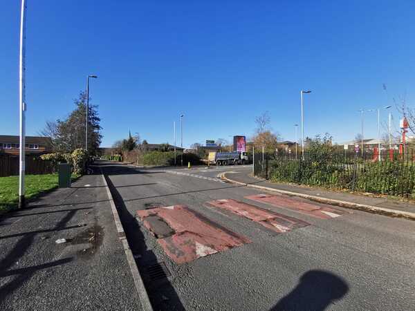 Looking down Goodiers Drive with red speed cushions in the middle of the road, pavement on the left and a fence on the right. Signs for Burger King are seen in the background.